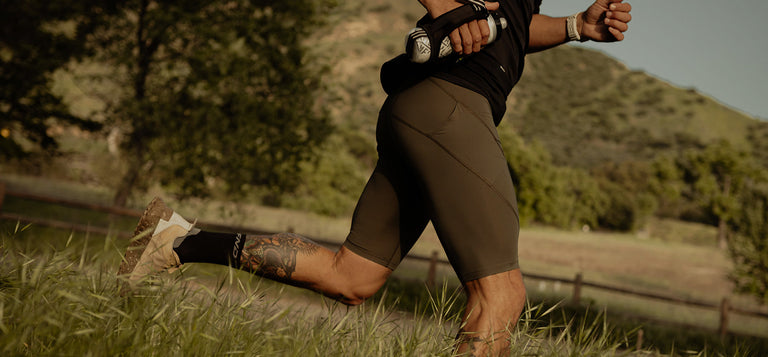 Person running outdoors on a grassy trail, wearing brown shorts, a black top, and athletic shoes, with mountains and trees in the background.