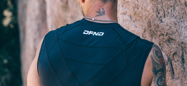 A person wearing a black sleeveless top with "DFND" printed on the back, showing a bird neck tattoo and a partial arm tattoo, standing near a stone wall.