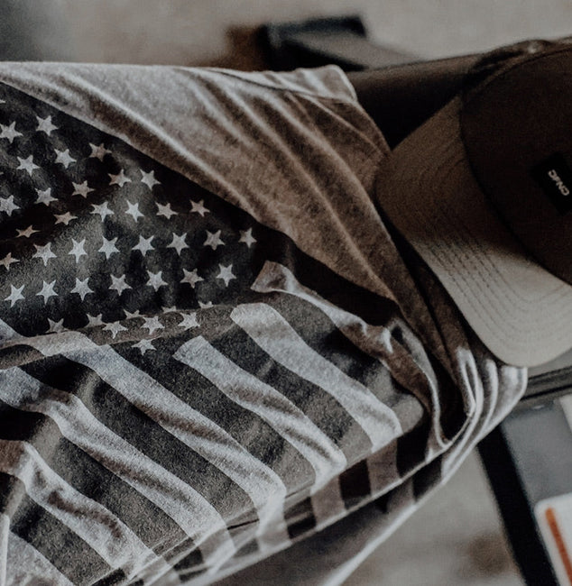 A gray t-shirt with a black-and-white American flag design and an olive green baseball cap are laid out on a gym bench.