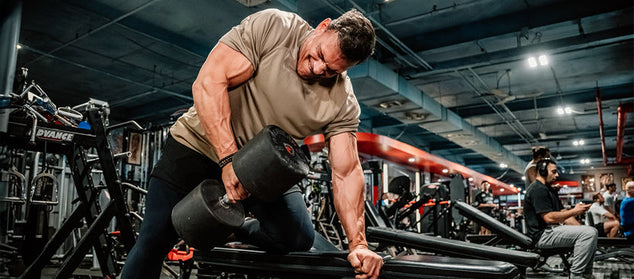 A man in a gym performs a one-arm dumbbell row on a bench, surrounded by gym equipment and other people working out.