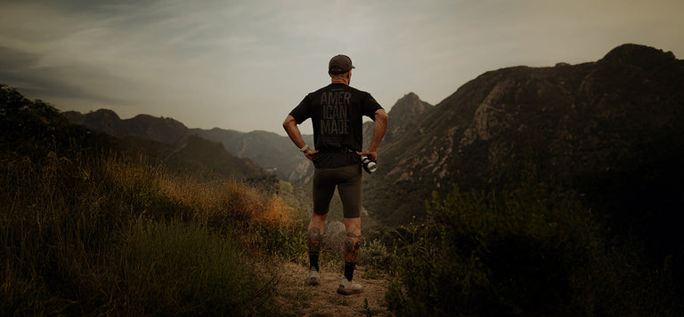 A person in athletic gear stands on a dirt trail, facing away, overlooking a mountain landscape under a cloudy sky.