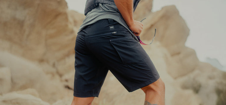Person wearing dark shorts and a gray shirt standing outdoors on rocky terrain, holding sunglasses in one hand.