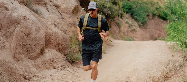 A man wearing a cap, black shirt, shorts, and a backpack runs on a dirt trail between rocky cliffs and greenery.