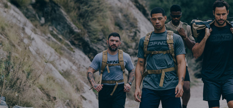 A man wearing a cap, black shirt, shorts, and a backpack runs on a dirt trail between rocky cliffs and greenery.