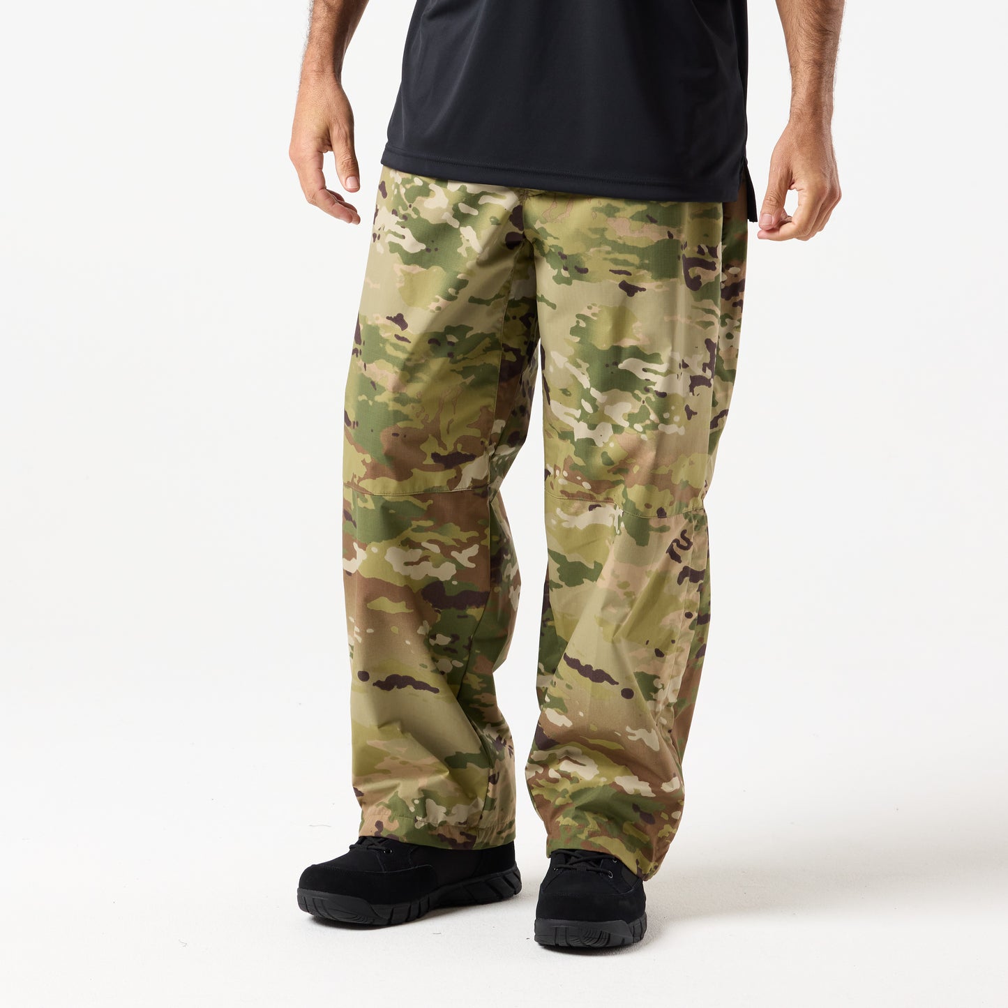 A person models loose-fitting, waterproof DFND Assault Pants in camouflage, paired with a black shirt and black shoes, standing against a plain white background.
