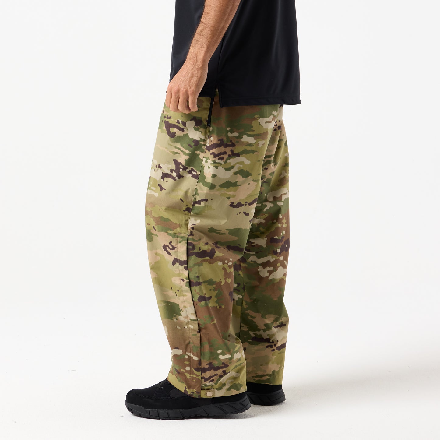 A person wearing loose-fitting DFND Assault Pants in camouflage, a black shirt, and black shoes stands against a plain white background.