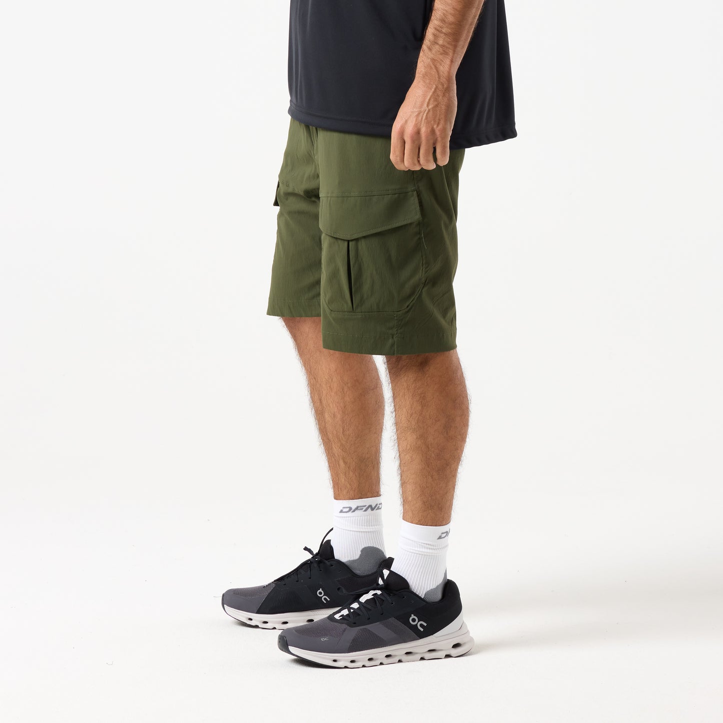 A person wears DFND MISSION CARGO SHORTS in olive green with zippered pockets, paired with black athletic shoes, white socks, and a dark shirt, all set against a plain white background.