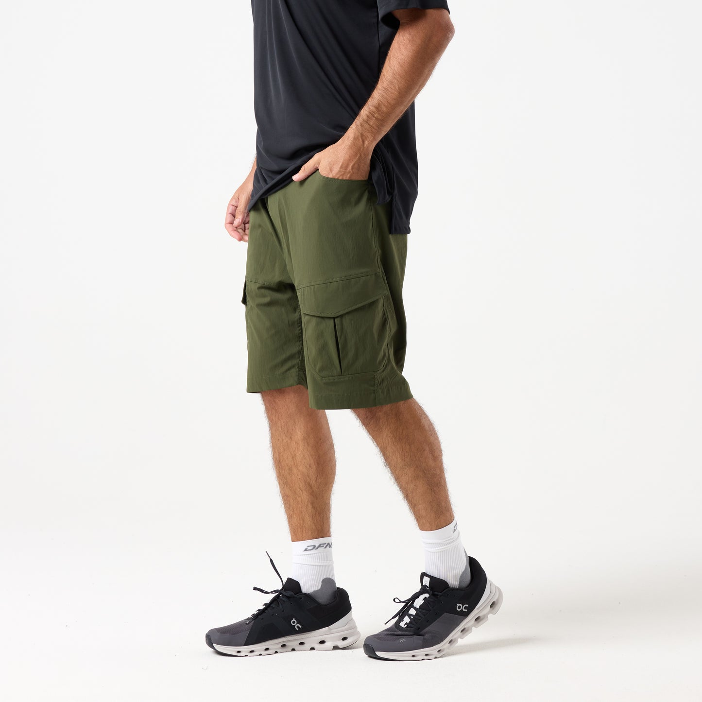 A person wearing DFND MISSION CARGO SHORTS in olive green, featuring zippered cargo pockets, along with a black shirt, white socks, and black athletic shoes stands against a plain white background.