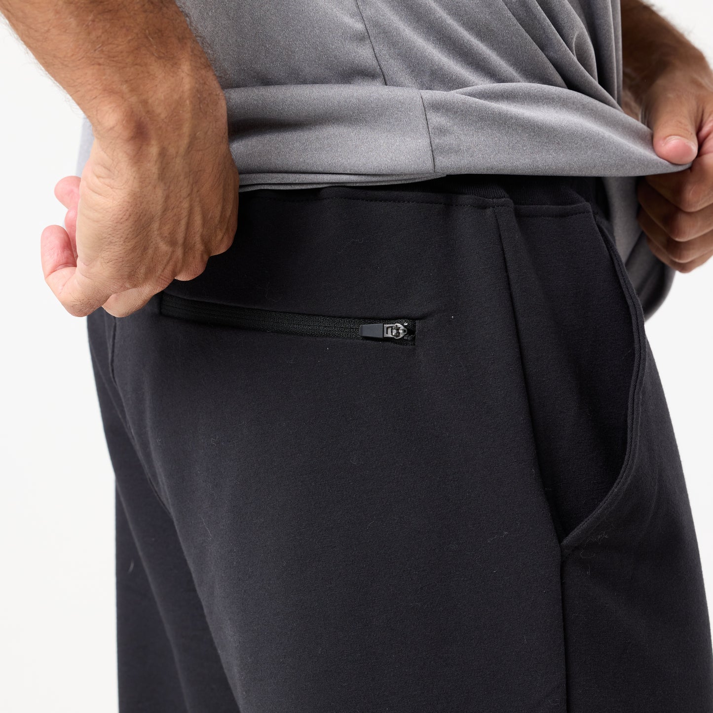 A person lifts a gray shirt, revealing the waistband and zippered back pocket of DFND’s IR Travel & Recover Rx Jogger in black, crafted with CELLIANT infrared-responsive textiles.