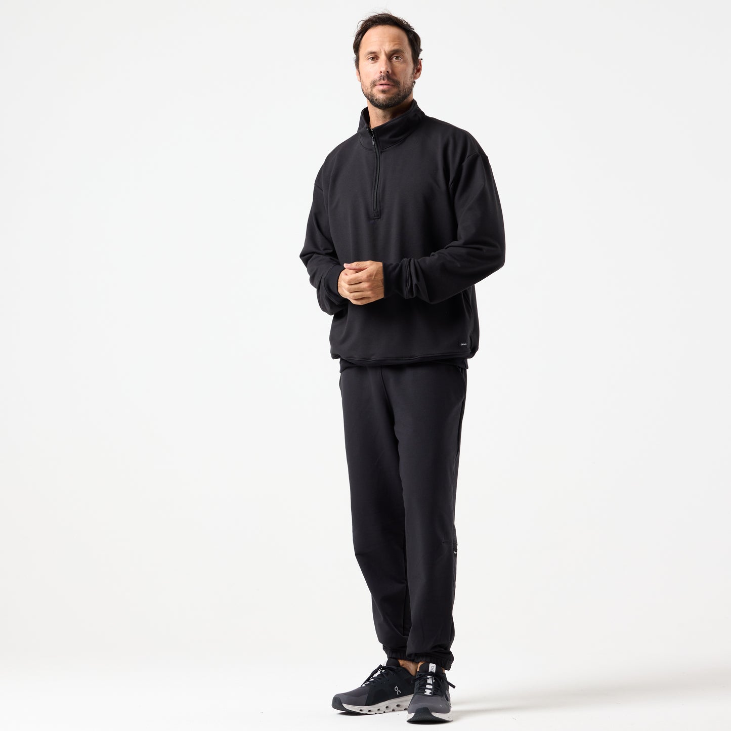 A man stands against a plain white background wearing the DFND IR Travel & Recover 1/4 ZIP, matching joggers, and black athletic shoes designed to enhance tissue oxygen.