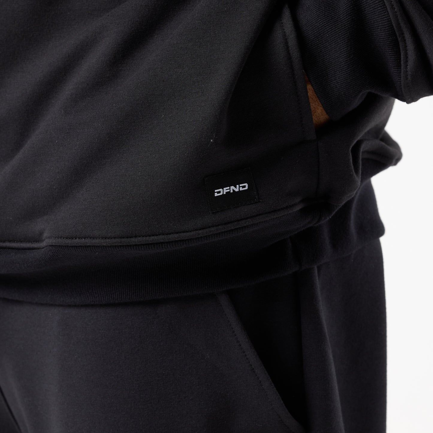 Close-up of a person wearing the DFND IR Travel & Recover 1/4 ZIP in black, made with CELLIANT textiles and featuring a subtle "DFND" logo on the sweatshirt, one hand partially in the pocket.