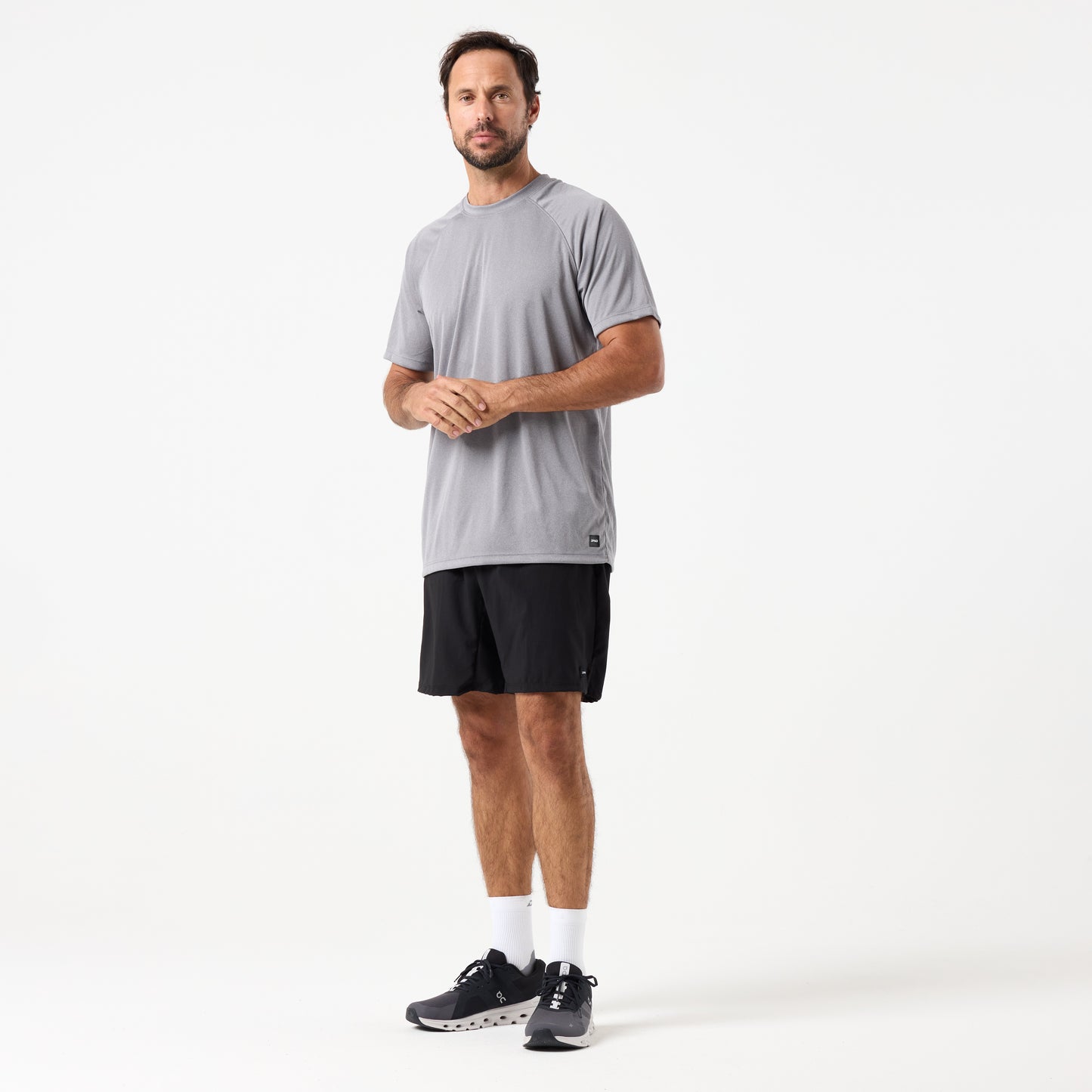A man stands facing forward against a white background, wearing DFND MALIBU 7" PT SHORTS in black, a gray short-sleeve shirt, white socks, and black athletic shoes.