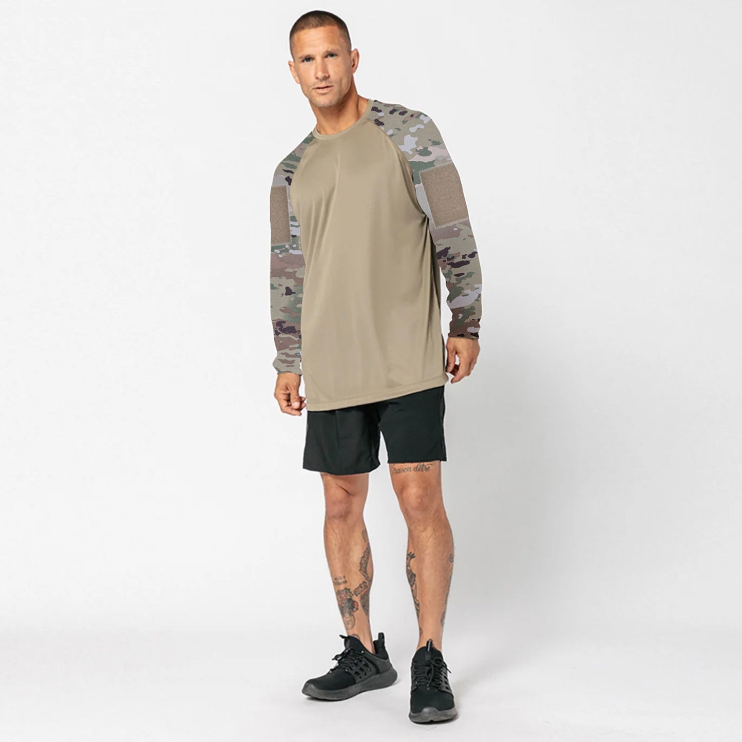 A man stands against a plain background wearing the DFND SPEED TECH LS SHIRT WITH VELCRO featuring tan fabric and camouflage sleeves, paired with black shorts and black athletic shoes.