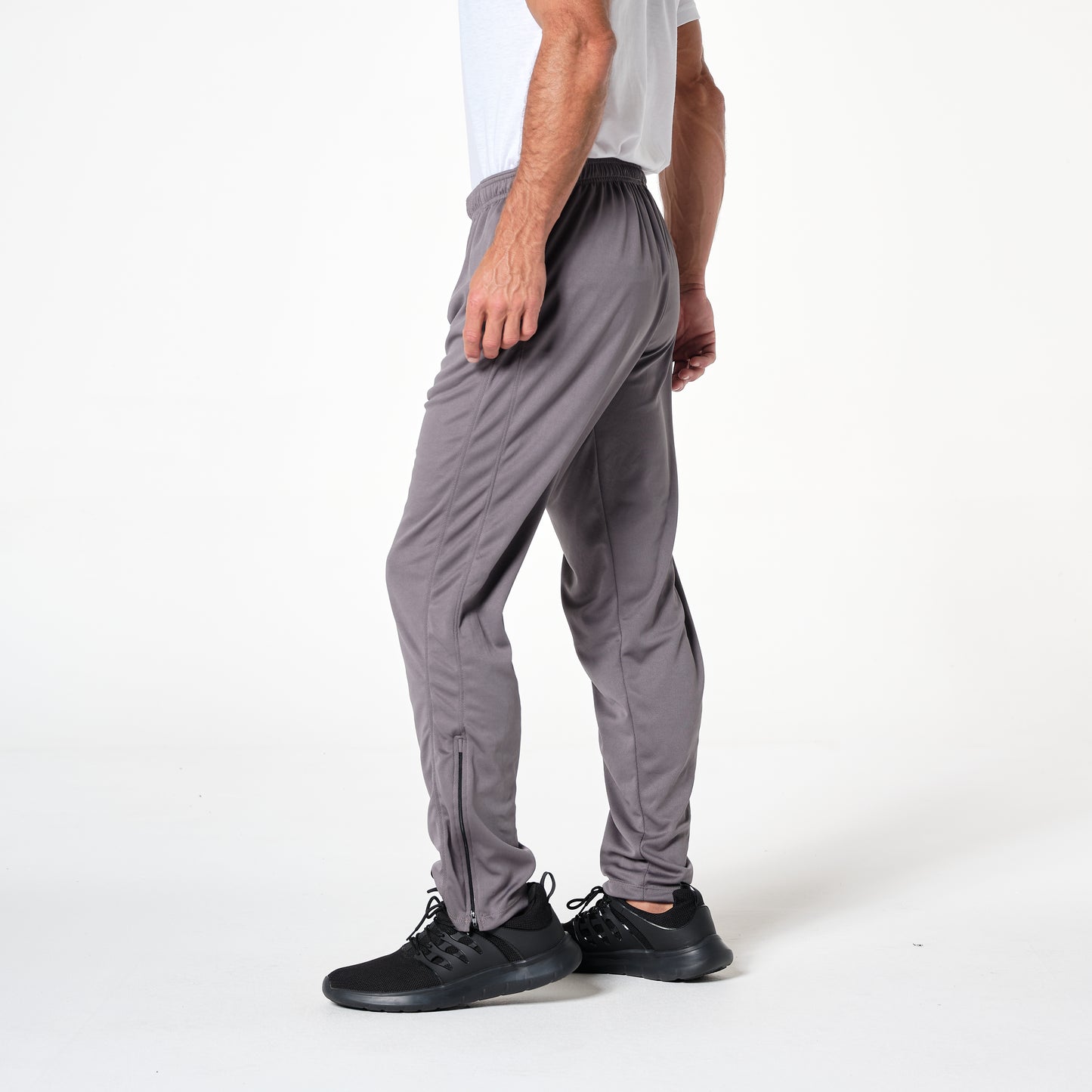 A man standing in gray athletic pants and a white shirt, facing slightly sideways. He is wearing black shoes. The background is plain white.