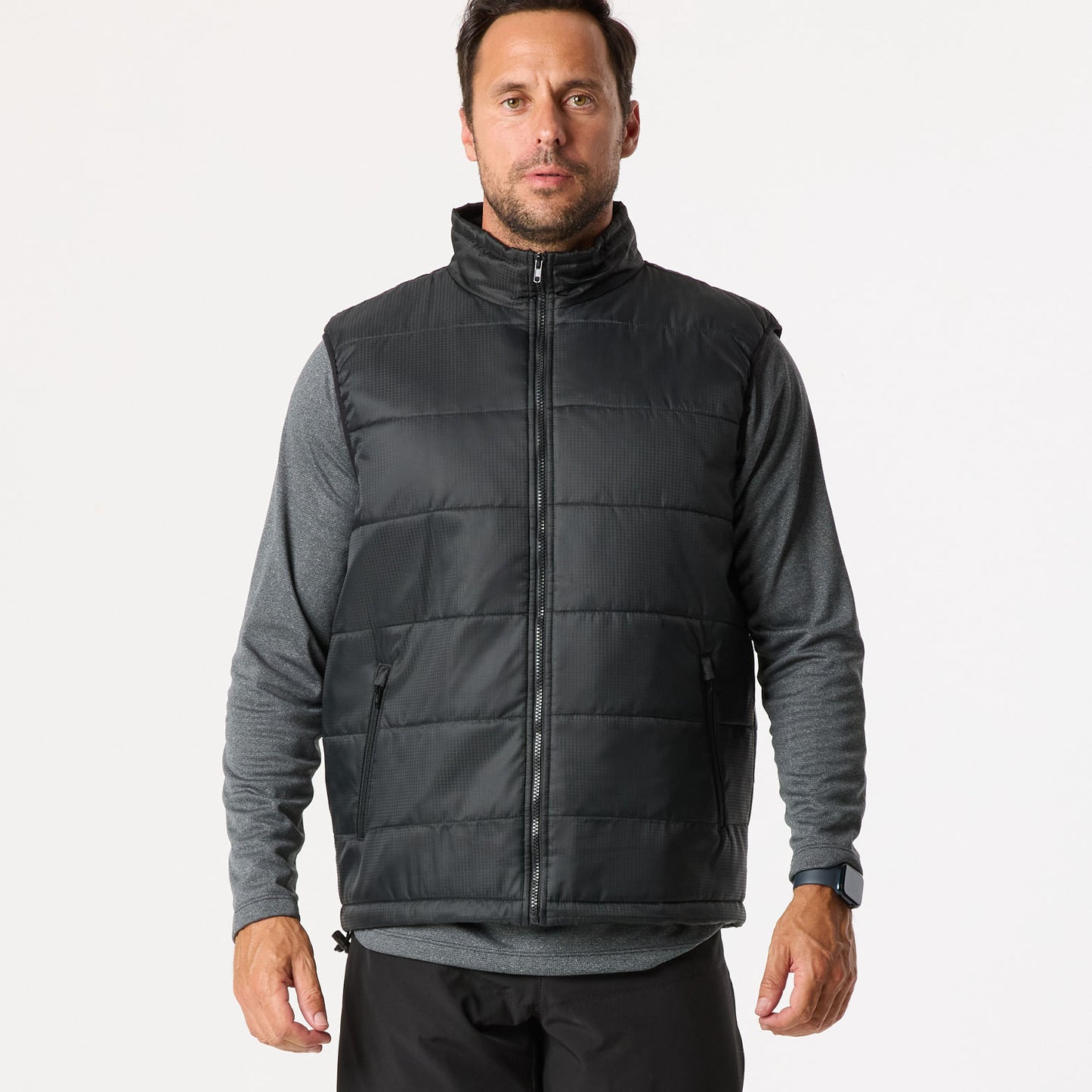 A man wearing a black quilted vest over a gray long-sleeve shirt stands against a plain background.