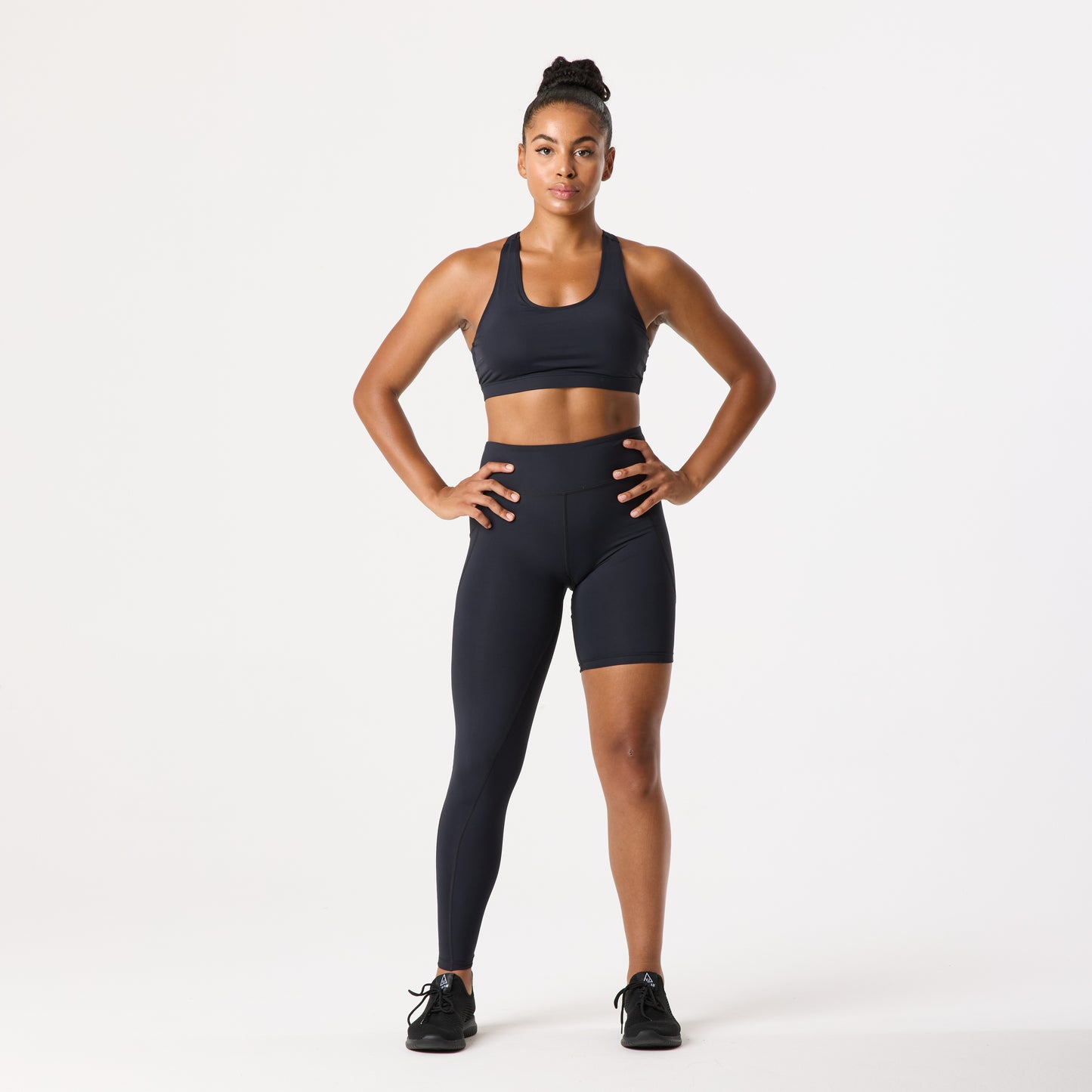 A person wearing a mismatched pair of leggings and shorts and a sports bra stands confidently with hands on hips against a plain background.