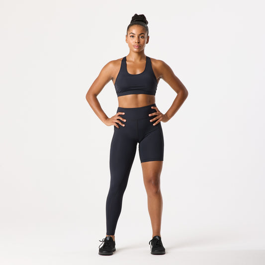 A person wearing a mismatched pair of leggings and shorts and a sports bra stands confidently with hands on hips against a plain background.
