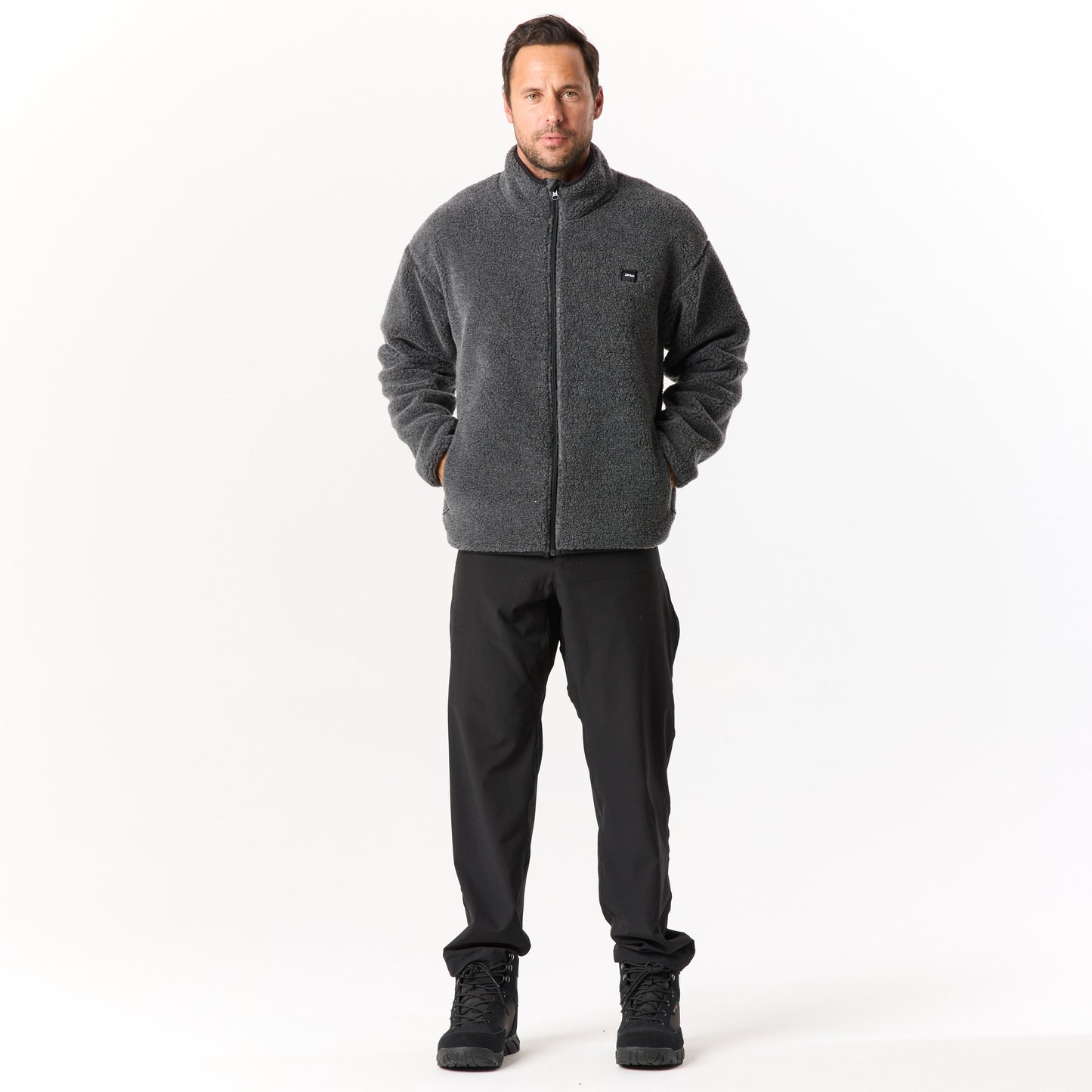 A person stands against a plain white background wearing a gray fleece jacket, black pants, and black shoes, hands in pockets.