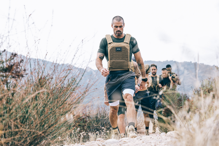 Four people hiking on a rocky trail wearing tactical vests and sportswear, with dry shrubs and mountains in the background.