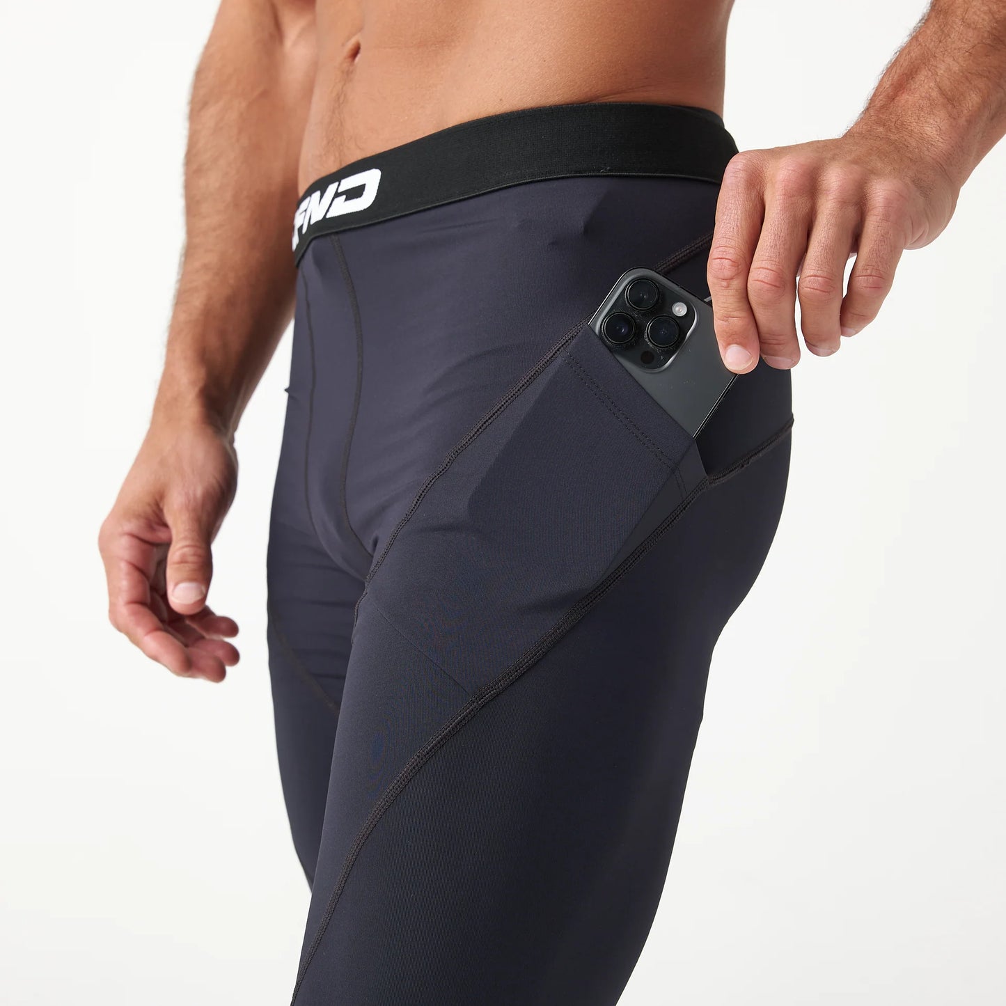 A person places a smartphone into the side pocket of black DFND Compression Shorts from the 3-Pack, featuring a branded waistband and showcasing stylish CELLIANT-powered infrared recovery sleepwear.