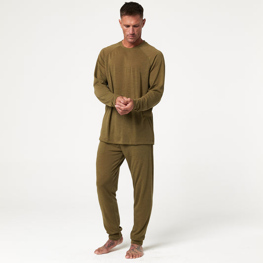 A person stands barefoot in olive green long-sleeve loungewear, clasping hands, against a plain white background.