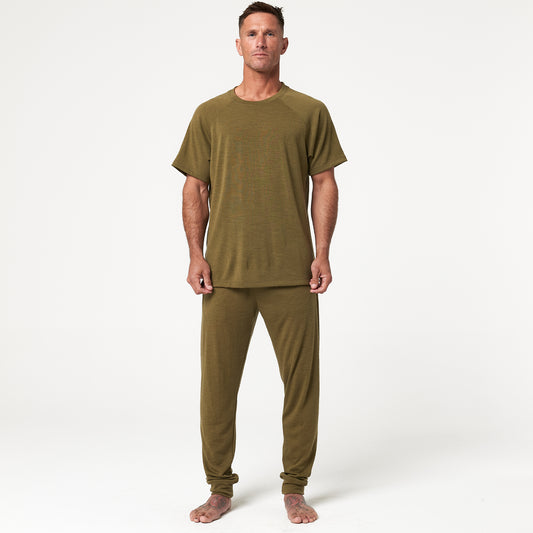 A person stands facing forward, wearing a matching olive green short-sleeve shirt and pants, barefoot, on a plain white background.