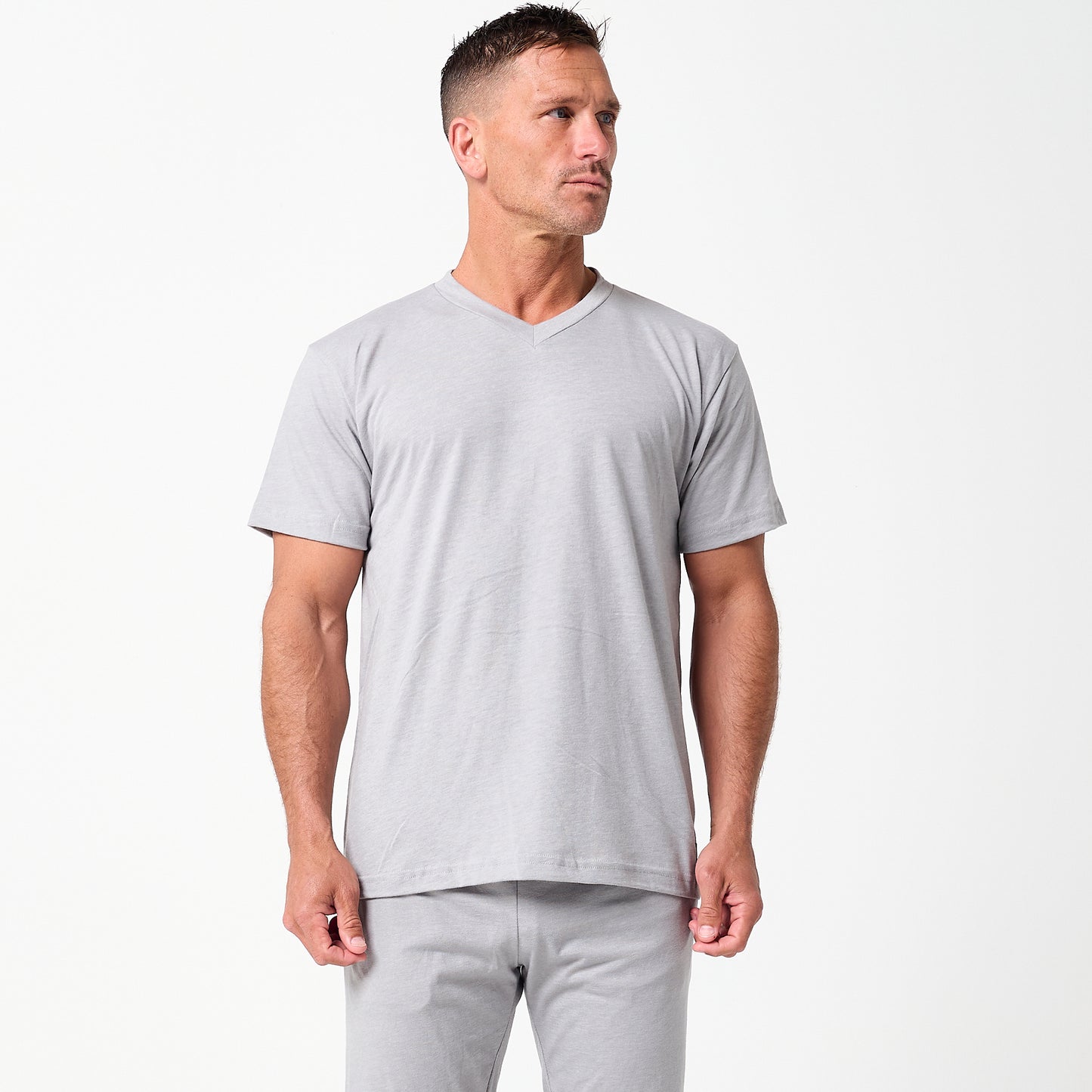 A man stands against a plain white background, wearing the DFND Cooling Recovery Sleepwear SS Shirt by DFND and matching gray pants, made from infrared-responsive textiles, looking slightly to the side.