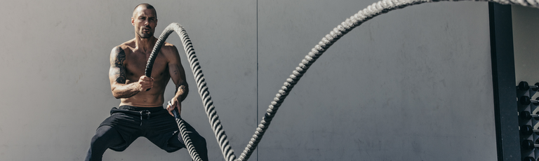 A shirtless man is exercising outdoors with battle ropes, standing in a wide stance against a gray wall.