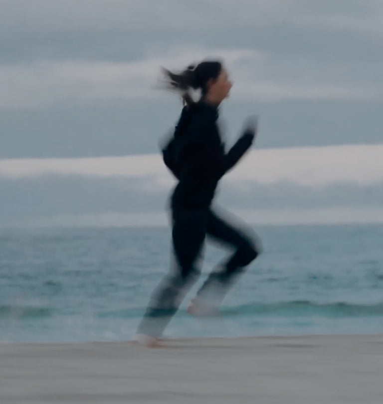 A person in dark clothing is running on a beach, with the ocean and a cloudy sky in the background.