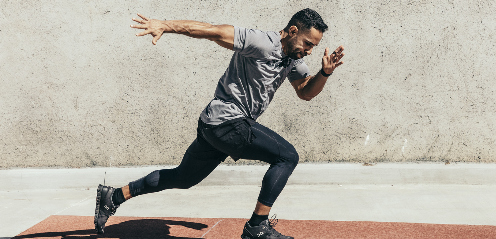 A man wearing a gray shirt and black pants is sprinting on a track against a concrete wall.