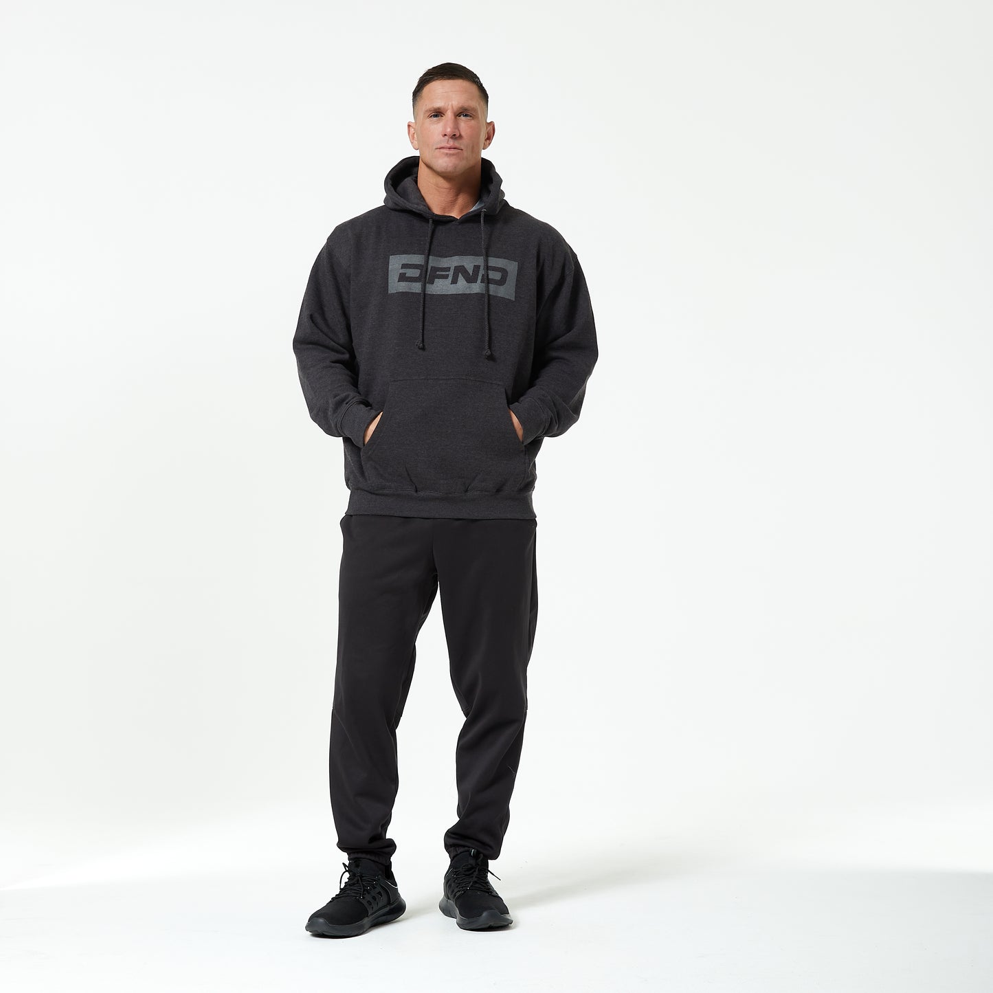 A man faces forward in a DFND TITAN LOGO FLEECE HOOD, dark joggers, and black sneakers against a plain white background—ideal for cold weather training.
