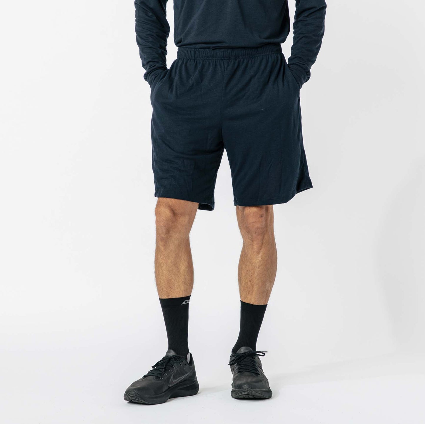 A person wearing black long-sleeve shirt, black shorts, black socks, and black shoes stands with hands in pockets against a plain background.