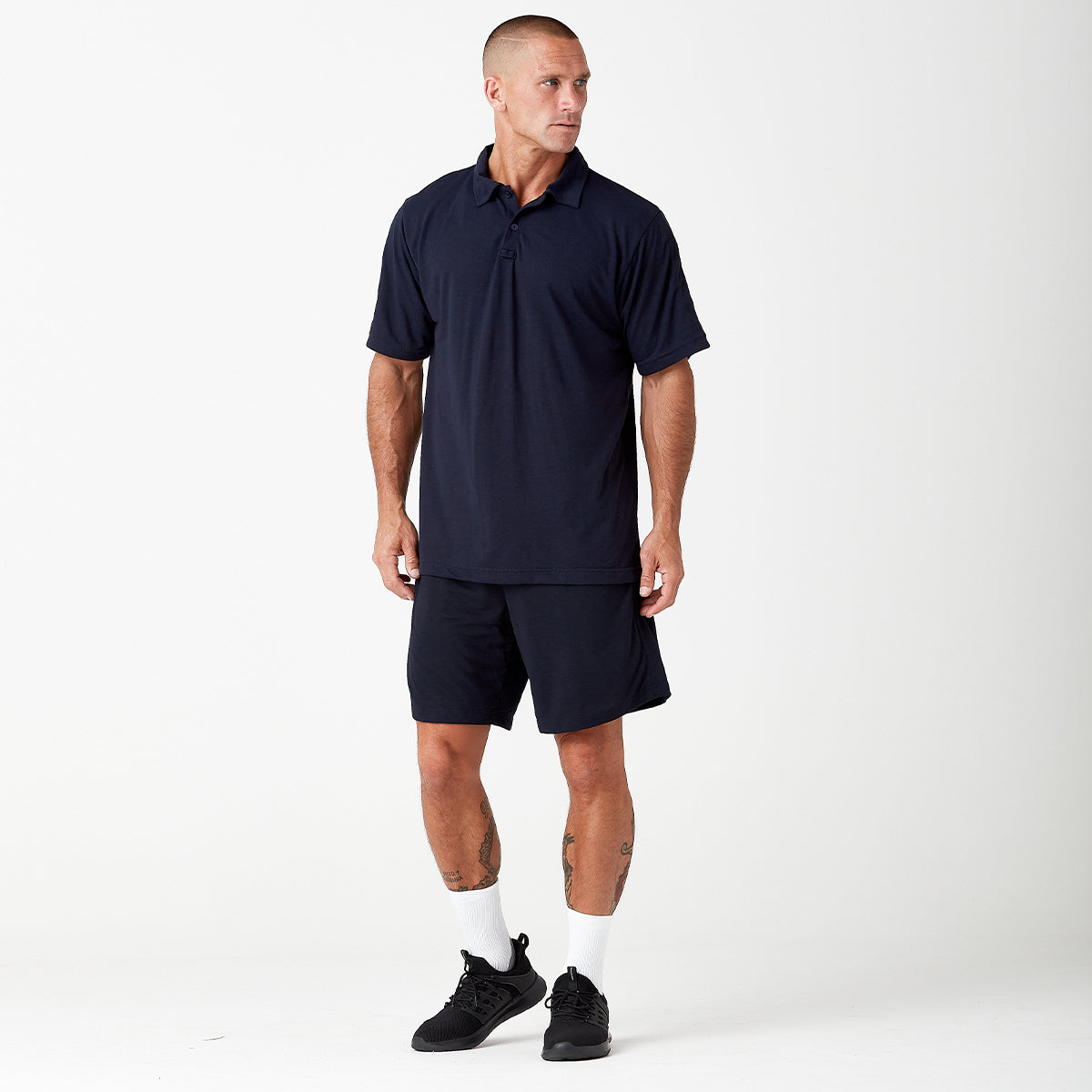 A person wearing a navy blue polo shirt, matching shorts, white socks, and black sneakers stands facing left against a plain white background.