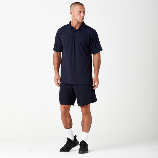 A person wearing a navy blue polo shirt, matching shorts, white socks, and black sneakers stands facing left against a plain white background.