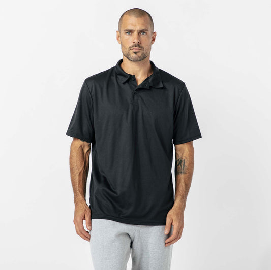 A man with a shaved head wearing a black polo shirt and gray pants stands against a plain white background.