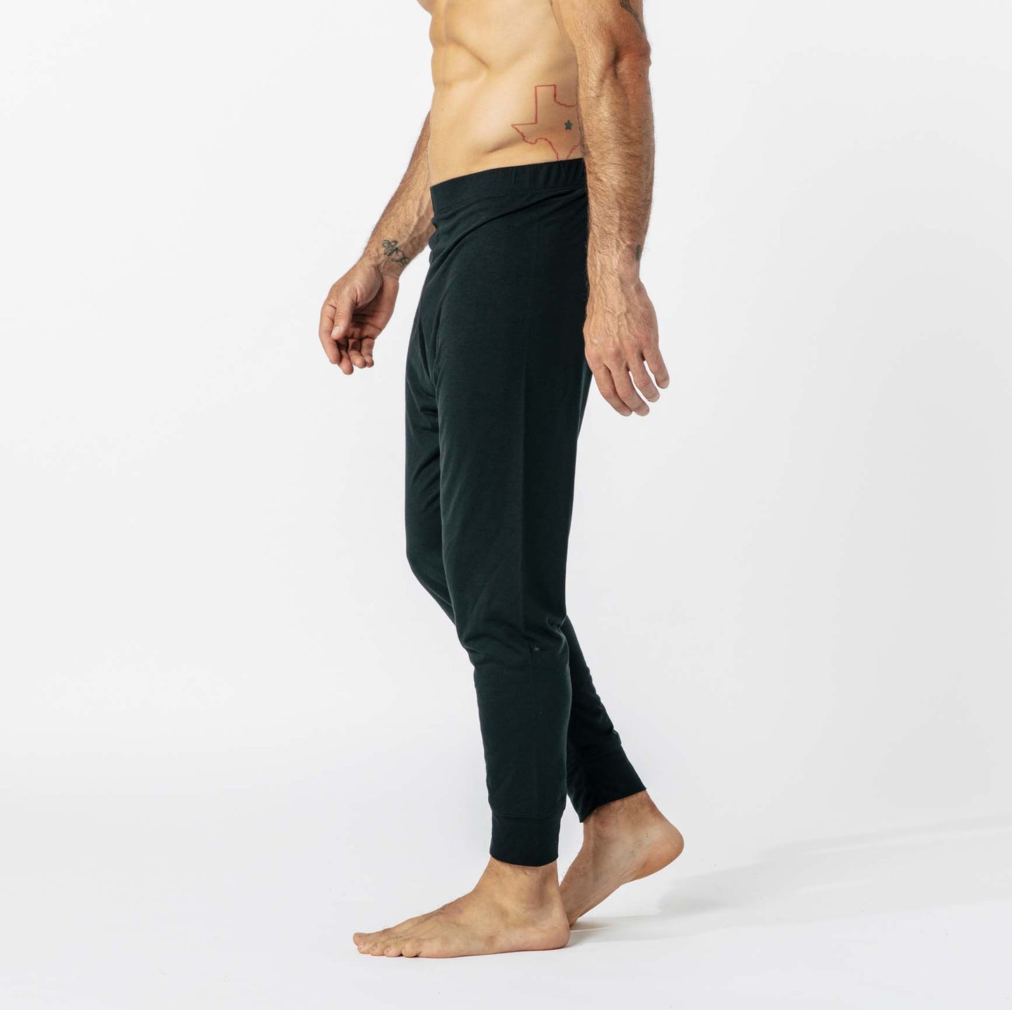 A shirtless person in dark joggers stands against a plain white background, facing sideways, with visible tattoos on the right arm and left abdomen.