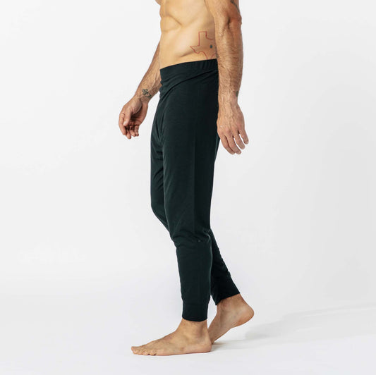 A shirtless person in dark joggers stands against a plain white background, facing sideways, with visible tattoos on the right arm and left abdomen.