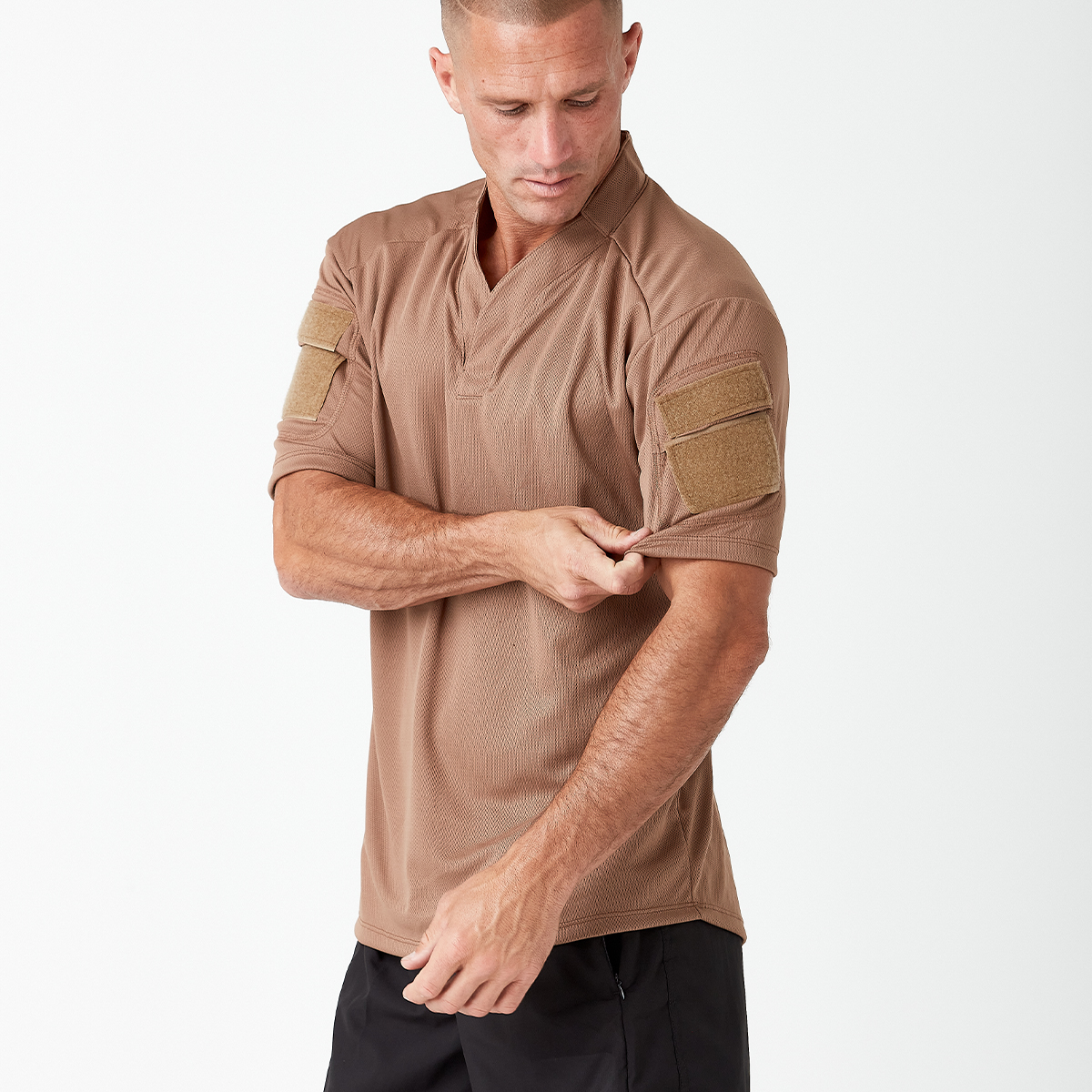 A man in a brown short-sleeve shirt adjusts his sleeve. The shirt has velcro patches on both arms. He is wearing black pants and is set against a plain white background.