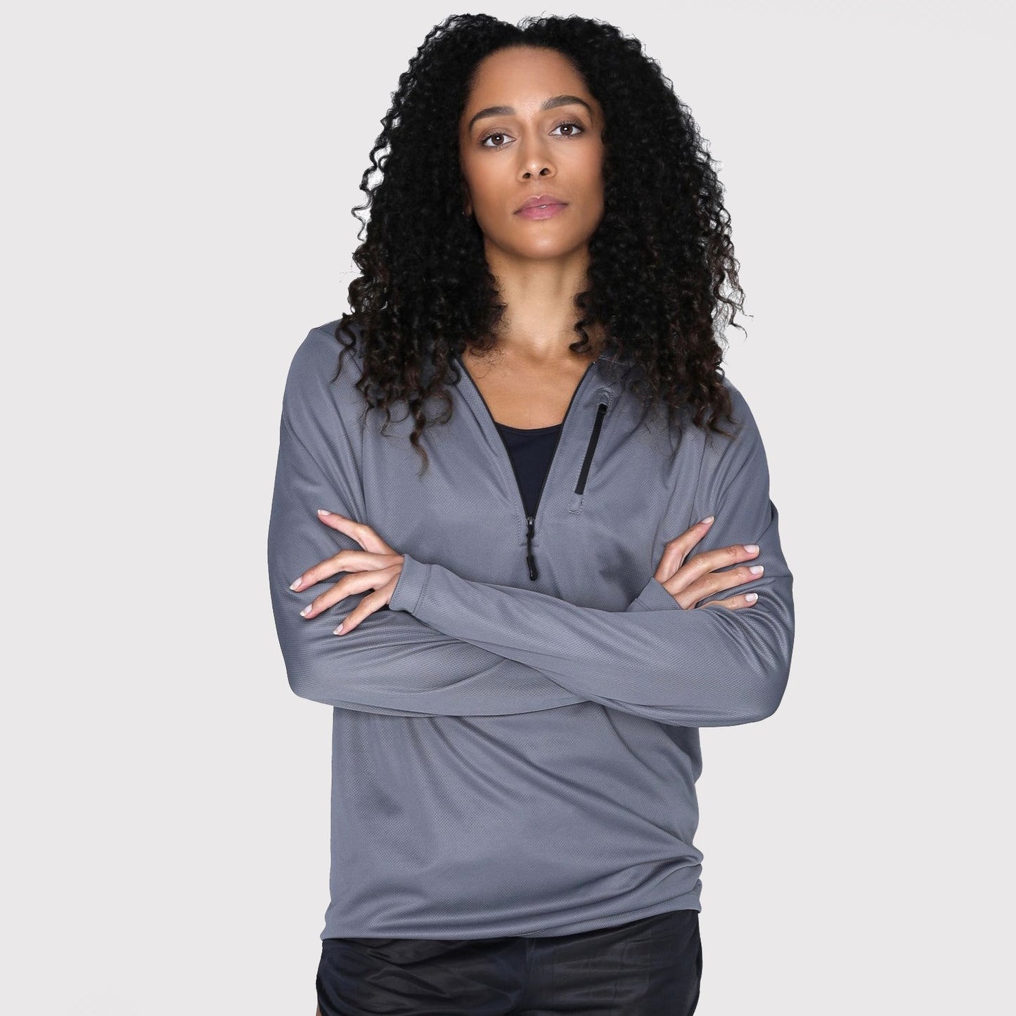 A woman with curly hair stands against a plain background, arms crossed, wearing the DFND SHIELD HOODED SUN SHIRT in gray over a black top, which features antimicrobial properties and SPF 50+ protection.