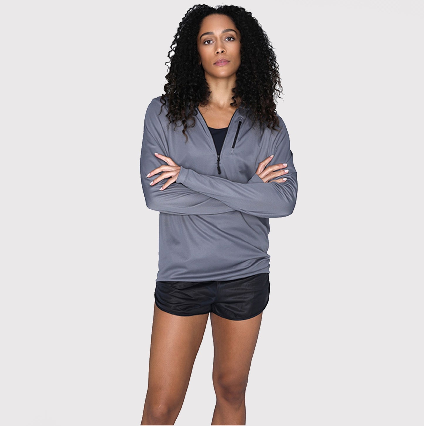 A person with curly hair stands against a plain background, wearing the DFND SHIELD HOODED SUN SHIRT with SPF 50+ protection and black shorts, arms crossed.