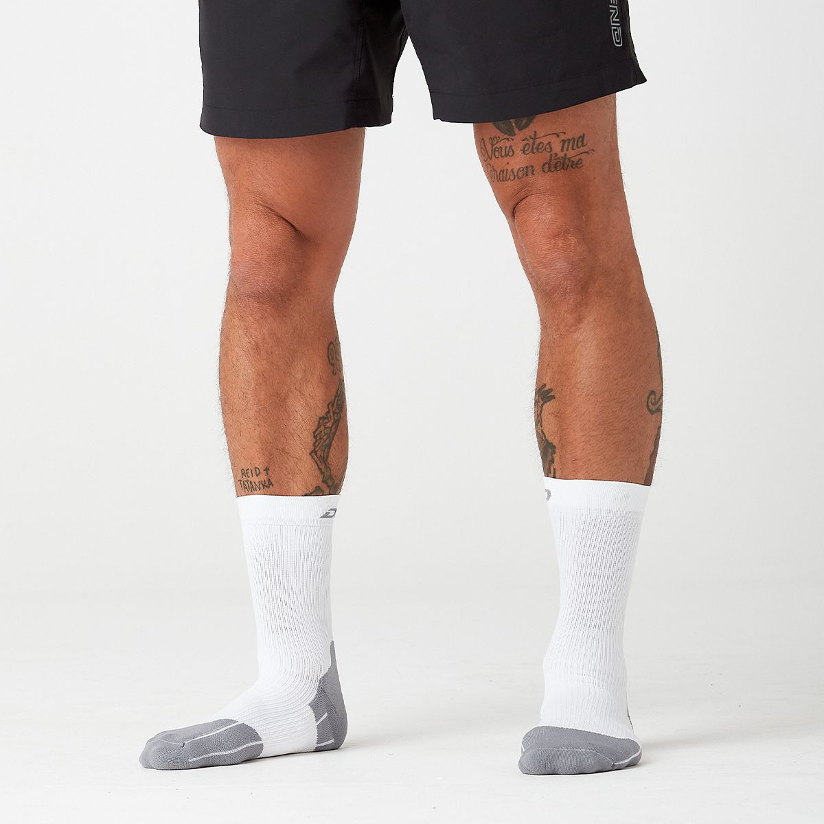 A person wearing black shorts and white socks with gray toes stands with legs slightly apart. Both legs have tattoos.