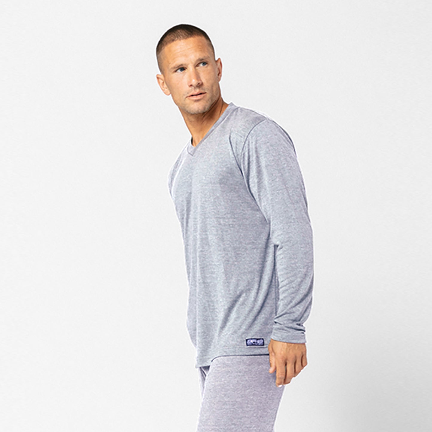 A man stands against a plain white background, looking to his left, wearing the DFND Cooling Recovery Sleepwear LS Shirt—a light gray long-sleeve shirt by DFND—designed to help improve sleep quality.