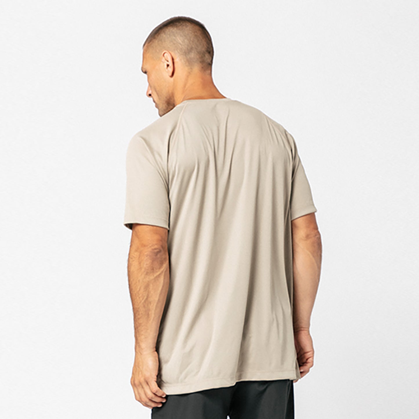 A man faces away, wearing a loose-fitting beige DFND SPEED TECH SS SHIRT - Raglan Sleeve with FUZE antimicrobial protection and dark pants against a plain light background.