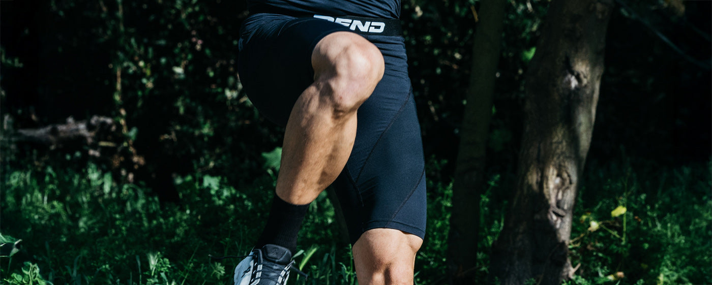 Person in black athletic shorts and a black shirt running outdoors, with one knee lifted and trees in the background.