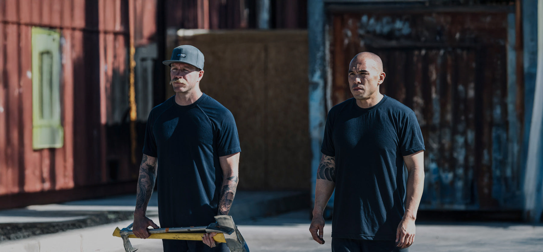 Two men with tattoos wearing dark T-shirts stand outdoors; one holds a sledgehammer and wears a cap, while the other looks ahead. Shipping containers are visible in the background.