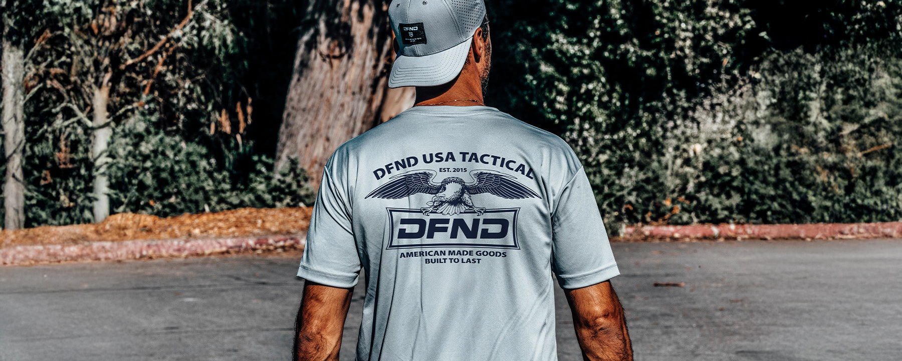 Man standing outdoors wearing a light gray "DFND USA Tactical" t-shirt and a backward cap, with trees and a paved ground in the background.