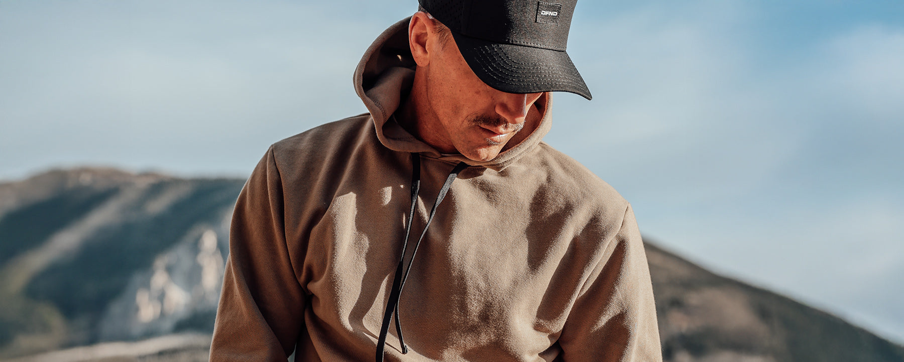 A person wearing a brown hoodie and black cap stands outdoors with mountains and sky in the background.
