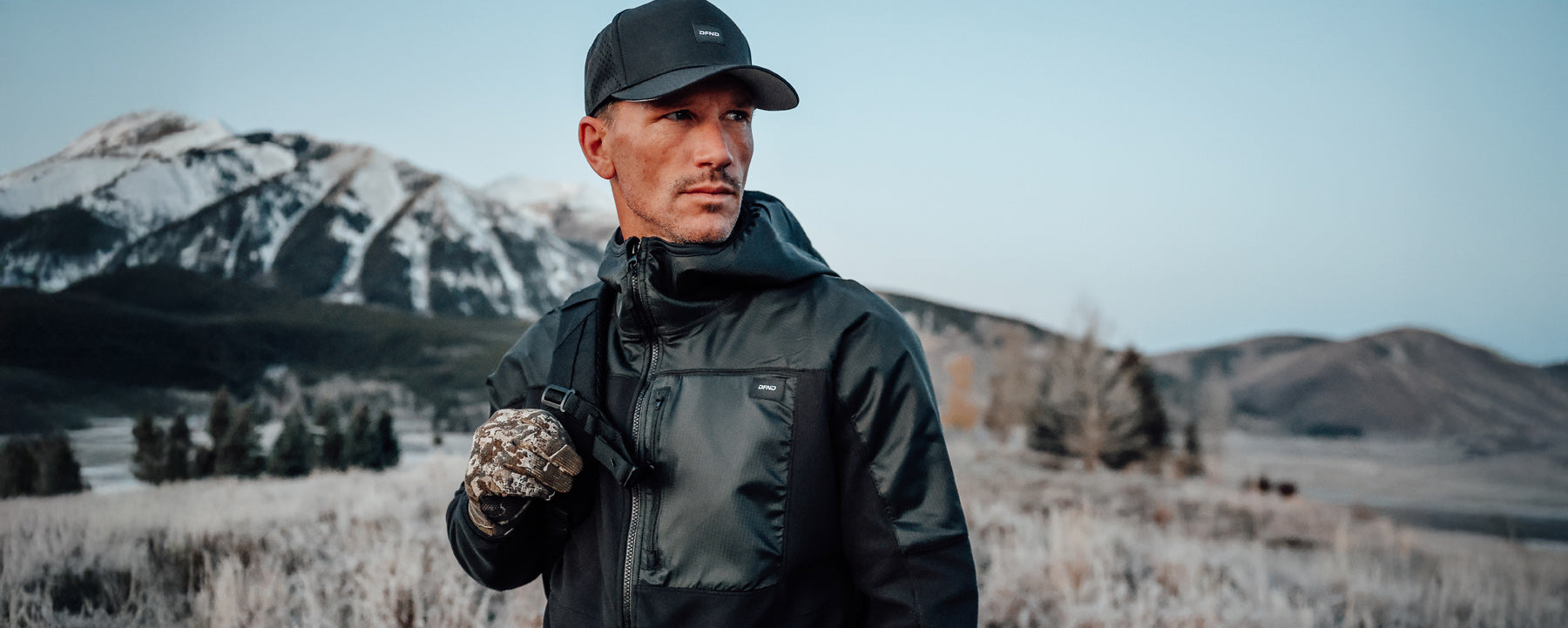 Man wearing a black jacket, cap, and camouflage glove stands outdoors with snow-covered mountains and trees in the background.