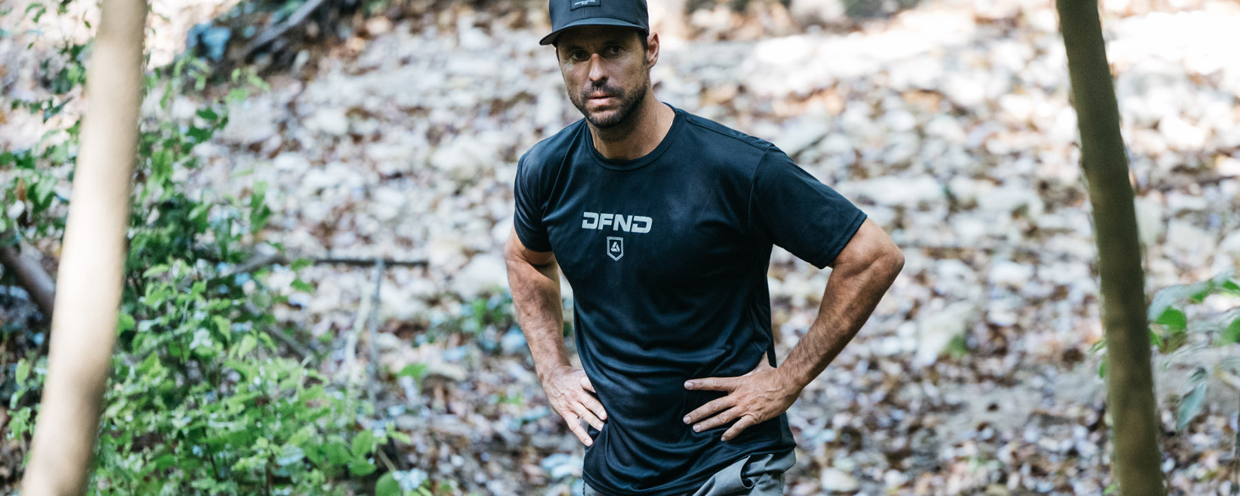 Man in a black DFND shirt and cap stands outdoors with hands on hips, surrounded by greenery and a rocky ground.