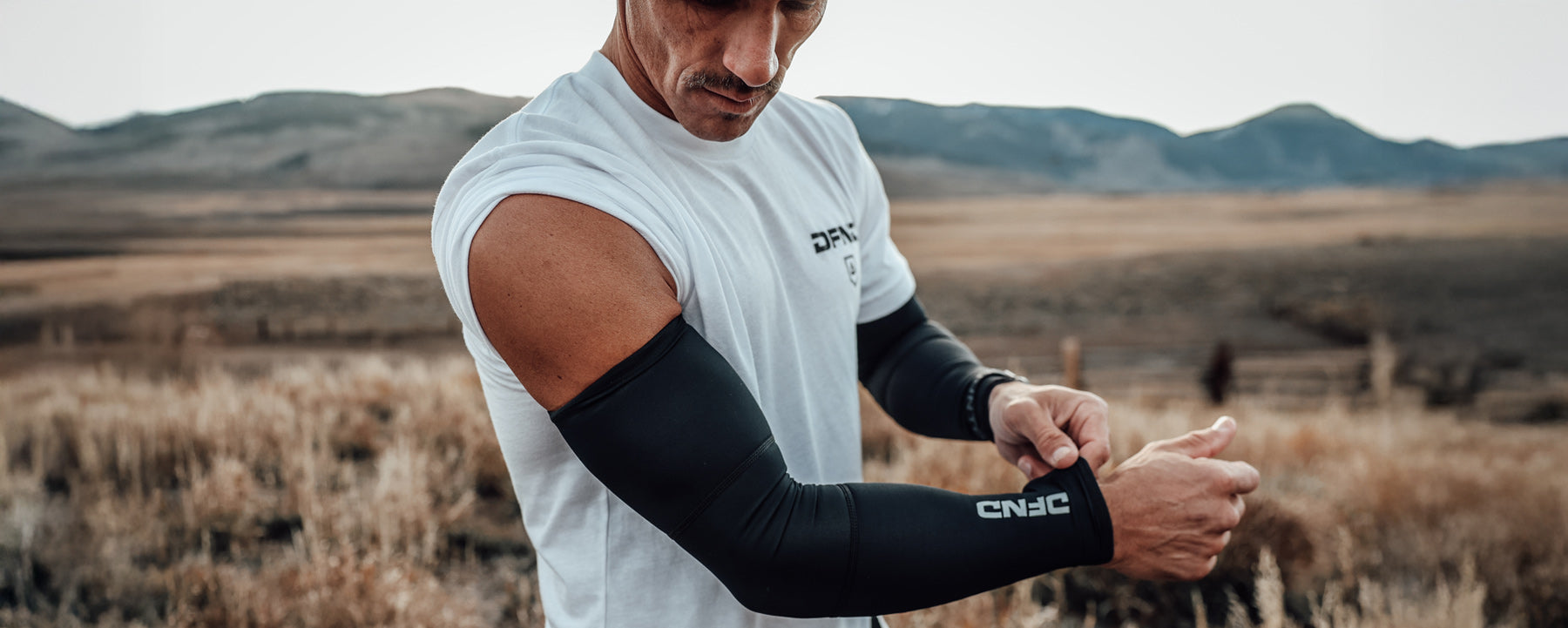 A person in a white t-shirt stands outdoors in a dry field, adjusting a black compression arm sleeve with "CNHC" written on it. Mountains are visible in the background.